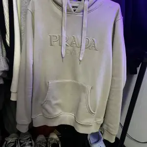 Säljer en snygg beige hoodie från Prada i storlek L. Den har en stor ficka fram och justerbar huva med dragsko. Perfekt för en avslappnad stil och passar till alla säsonger. Märkesloggan är broderad på framsidan för en lyxig touch.