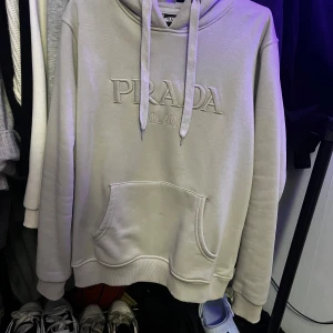 Beige hoodie från Prada inte äkta! - Säljer en snygg beige hoodie från Prada i storlek L. Den har en stor ficka fram och justerbar huva med dragsko. Perfekt för en avslappnad stil och passar till alla säsonger. Märkesloggan är broderad på framsidan för en lyxig touch.