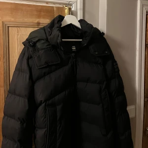 Svart dunjacka från G-Star - Tjaaaaaaaa, Säljer en svart dunjacka från G-Star i mycket bra skick. Jackan har en pufferdesign med dragkedja och knappar, samt praktiska fickor. Perfekt för kalla vinterdagar. Den är långärmad och har en stilren look som passar till det mesta. Bara att skriva för fler bilder. Inga defekter eller skador, priset är ej hugget i sten🤗Ny pris runt 2500!