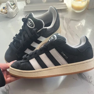 Svarta Adidas Campus sneakers - Säljer ett par klassiska svarta Adidas Campus! Är i väldigt bra skick och har bara en liten skråma som inte syns när man använder dem (se sista bilden) Nypris 1500kr kommer med vita snören om man vill, men de är väldigt smutsiga. Finns ju nya att köpa online.