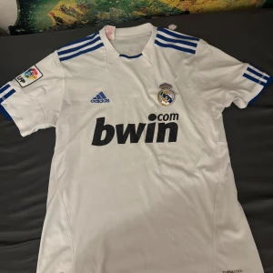 Vit Real Madrid fotbollströja från Adidas - Säljer en vit Real Madrid fotbollströja i bra skick. Den har korta ärmar och blå detaljer på axlarna. Tröjan är från Adidas och har klubbens emblem på bröstet samt sponsorloggan 'bwin.com'. Perfekt för alla fans av laget!