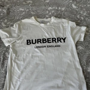 Vit t-shirt från Burberry - Säljer en snygg vit t-shirt från Burberry med svart text 'BURBERRY LONDON ENGLAND' på framsidan. Perfekt för en stilren och klassisk look. T-shirten är i bra skick och passar till både vardag och fest. Skicka ett meddelande vid intresse!