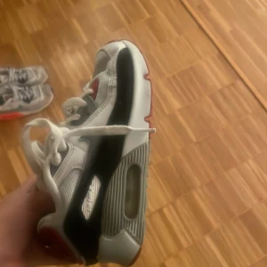 Nike Air Max i vitt, svart och rött - Säljer ett par Nike Air Max sneakers i vitt, svart och rött. De har en klassisk design med synlig luftdämpning i sulan och snörning framtill. Perfekta för både vardag och träning. Skorna är i bra skick med lite slitage på sulan.