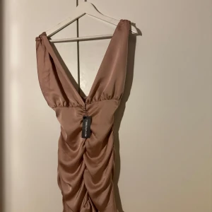Champagnefärgad klänning från PrettyLittleThing - Säljer en elegant champagnefärgad klänning från PrettyLittleThing. Den har en djup v-ringning och är ärmlös, perfekt för festliga tillfällen. Klänningen har en snygg rynkad detalj framtill som ger en smickrande passform. Materialet känns lyxigt och är i mycket bra skick. Perfekt för nyår eller en utekväll! ✨