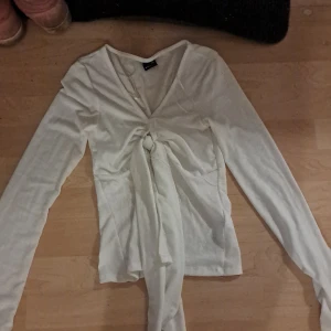 Vit långärmad topp från Gina Tricot - Säljer en vit långärmad topp från Gina Tricot i storlek XXS. Toppen har en snygg v-ringning och knytning framtill som ger en fin detalj. Perfekt för både vardag och fest! Har använt en gång men säljer pgd för liten obs köpte för 400