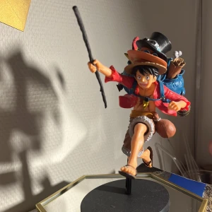 Figur av karaktär med hatt och stav - Monkey D. Luffy figur. Pinnen går att ta bort och sitter inte fast. Mycket fina detaljer från de olika arken. Även alla 3 brödernas hattar på. Figuren står på ett svart stativ och ser ut att vara i rörelse, vilket ger en dynamisk känsla. Perfekt för fans av äventyr och samlarobjekt!