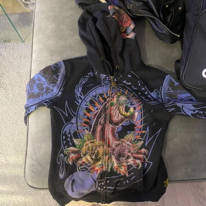 Ed hardy Svart hoodie med dödskallemotiv - Cool svart hoodie med färgglatt dödskallemotiv och spelkort. Den har en dragkedja framtill och huva. Ärmarna har detaljer med rosor och fåglar i blått och rött. Perfekt för en unik och edgy stil!