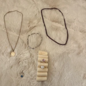 Smyckeset med halsbannd armband och ringar  - Säljer ett fint smyckeset med olika delar. Ett guldigt halsband med 'BEST' och tre små stenar, ett silverfärgat hjärthänge med blå detalj, och två ringar - en med rosa hjärta och en med glittriga stenar. 