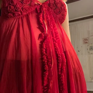 Röd klänning med rosett och volangdetaljer - Säljer en superfin röd klänning med volangdetaljer och en söt rosett framtill. Den har en romantisk stil med blommiga detaljer på bysten och är perfekt för festliga tillfällen. Klänningen är axelbandslös och har en luftig passform.