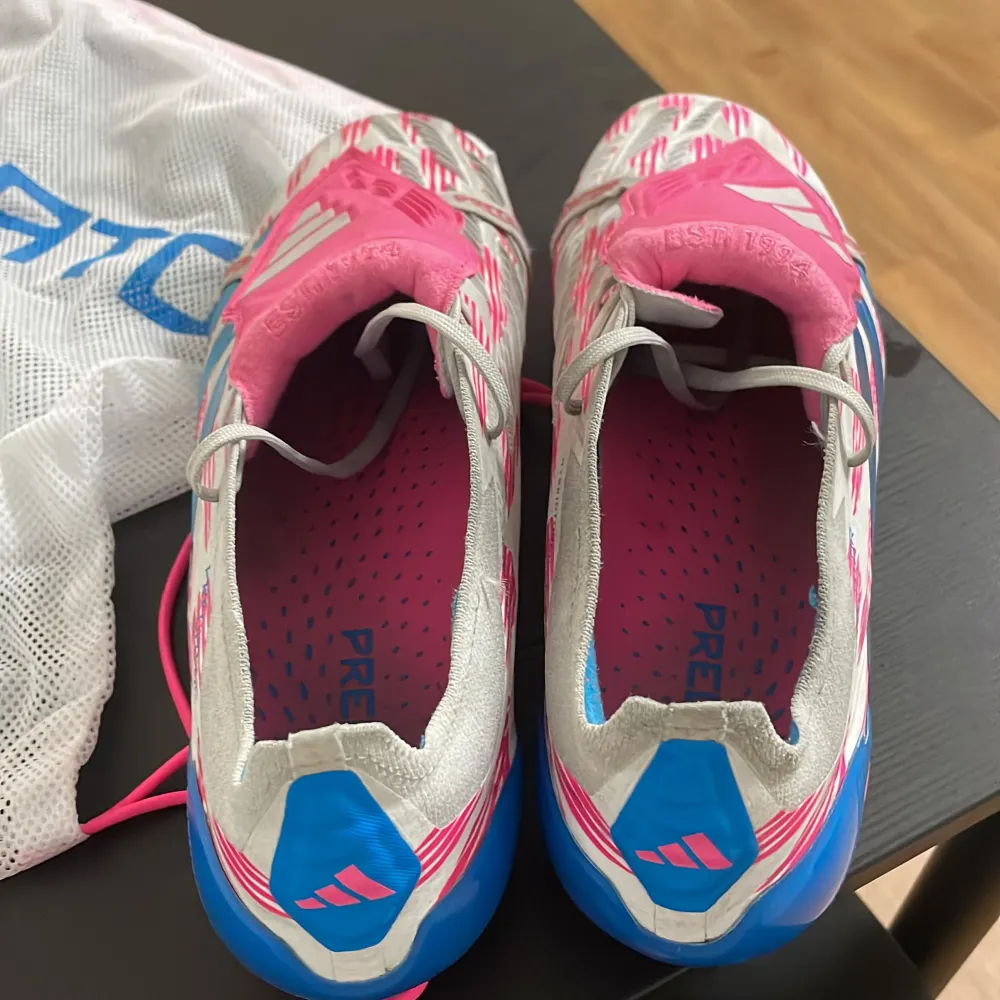 Säljer ett par Adidas Predator Elite fotbollsskor i vitt, rosa och blått. Skorna har en cool design med rosa och vita mönster och blå sula. De är i mycket bra skick då de inte används på mer än 3 veckor och ca 8 träningar och kommer med en praktisk förvaringspåse. Förvaringspåsen är lite sönder då ett av banden som håller upp skorna på ryggen är av. . Skor.