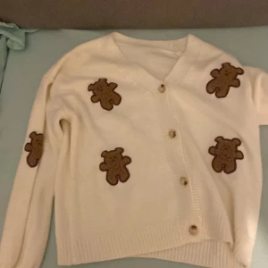 Vit kofta med nallemotiv - Supergullig vit kofta med bruna nallemotiv över hela framsidan. Den har en v-ringning och knappar framtill. Perfekt för att mysa till det under hösten eller vintern. Koftan är i bra skick och passar perfekt för en avslappnad stil.