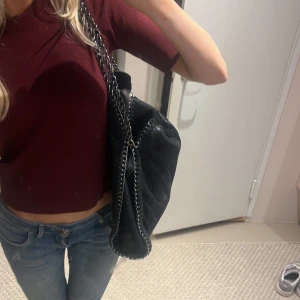 Svart axelväska med kedjedetaljer - Snygg svart axelväska i mycket bra skick med coola kedjedetaljer. Perfekt för både vardag och fest. Väskan är rymlig och Harlan även användas som skolväska dator ryms bra och en stilren design som passar till allt. Ett måste för garderoben!