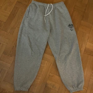 Grå mjukisbyxor från Nike x Stüssy - Säljer ett par grå mjukisbyxor från Nike i samarbete med Stüssy. Byxorna har en avslappnad passform med resår i midjan och muddar vid bensluten. De har en snygg logga på benet och är perfekta för chill dagar. Superbekväma och stilrena!