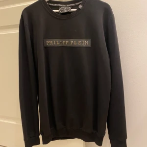 Philipp Plein Sweatshirt/Långärmad Tröja  - Säljer en stilren svart sweatshirt från Philipp Plein. Tröjan har en klassisk rund halsringning och ett tryck med märkesnamnet på bröstet. Perfekt för både vardag och festliga tillfällen. Den är i nyskick (oanvänd) och redo för en ny ägare!