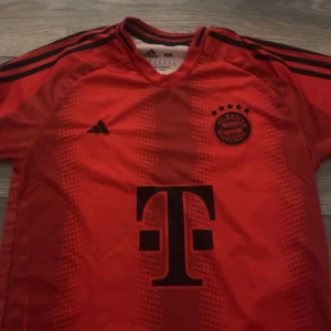 Röd FC Bayern München fotbollströja - Säljer en röd FC Bayern München fotbollströja i mycket bra skick. Tröjan har korta ärmar och är prydd med klubbens emblem och sponsorlogga på framsidan. På baksidan finns spelarnamnet 'Kane' och nummer 9.