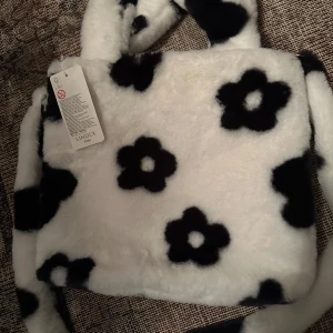Vit och svart blommig väska från Lindex - Superfin och fluffig väska från Lindex i vitt med svarta blommor. Perfekt för att ge en lekfull touch till din outfit! Väskan är i syntetmaterial och har en mjuk och gosig känsla. Den är idealisk för både vardag och festliga tillfällen.
