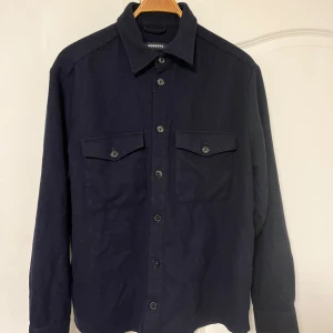 Mörkblå jacka/overshirt i ull från J.Lindeberg - Säljer en mörkblå jacka/overshirt från J.Lindeberg i ull. Perfekt för höst och vinter med sina långa ärmar och knappar framtill. Den har två bröstfickor med knappar som ger en snygg detalj. Passar både till vardags och fest. Orginalpris ligger på 2600 se bild 3. Jackan är blå som på de två första bilderna de andra är för att visa