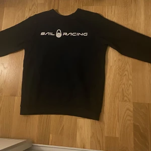 Svart tröja från Sail Racing - Säljer en svart tröja från Sail Racing i bra skick. Den har en vit logga på bröstet och en mindre logga på baksidan. Perfekt för en casual look eller en dag på stan. Tröjan är bekväm och stilren, ett måste i garderoben!