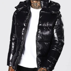 Svart pufferjacka med huva - Säljer en snygg svart pufferjacka i glansigt material. Den har en dragkedja framtill och en praktisk huva. Perfekt för kalla dagar och håller dig varm och stilren. Jackan är långärmad och har en modern look.