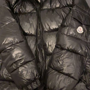 Svart pufferjacka från Moncler - Säljer en snygg svart pufferjacka från Moncler i mycket bra skick. Jackan är perfekt för vintern med sitt blanka polyester-material och har en dragkedja framtill. Den är långärmad och har en cool Moncler-logga på ärmen. Perfekt för att hålla sig varm och stilren under kalla dagar! 🖤