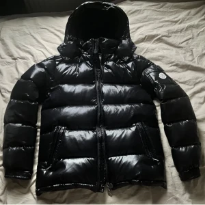 Moncler Maya  - Vid snabb affär (SÄNKER PRISET).                                   Säljer en snygg svart pufferjacka från Moncler i mycket bra skick. Jackan har en glansig finish och är perfekt för kalla vinterdagar. Den har en dragkedja framtill och en huva för extra värme. Moncler-loggan syns tydligt på ärmen. Perfekt för dig som vill hålla stilen även när det är kallt ute!
