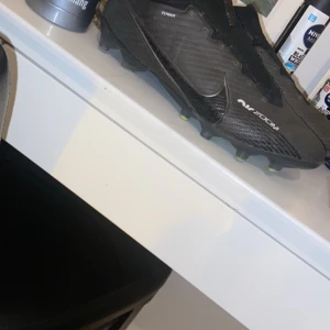 Svarta Nike fotbollsskor - Säljer ett par svarta Nike fotbollsskor i bra skick. Skorna har en snygg design med snörning och är perfekta för fotbollsträning. De har en slitstark sula med dobbar för bra grepp på planen. Perfekta för dig som vill ha stil och funktion i ett!