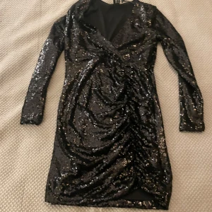 Svart paljettklänning från Vero Moda - Snygg svart klänning från Vero Moda i storlek XS. Den är täckt med glittriga paljetter och har en omlott-design framtill. Klänningen har långa ärmar och en tight passform, perfekt för festliga tillfällen. Den är i mycket bra skick och redo för nyårsfesten!