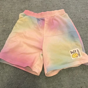 Färgglada shorts från SmileyWorld x H&M - Säljer ett par supercoola shorts från SmileyWorld x H&M. De har en härlig tie-dye effekt i pastellfärger som rosa, blå och gul. Perfekta för sommaren med en bekväm elastisk midja. Detaljerad med en söt smiley-patch som ger en extra touch av glädje. Perfekta för en avslappnad dag i solen!