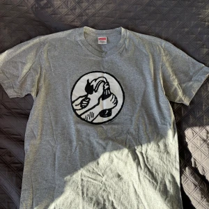 Grå t-shirt från Supreme - Säljer en grå t-shirt från Supreme i bomull och polyester. Den har ett coolt tryck på framsidan med en unik design. Perfekt för vardagsbruk eller att styla upp med en jacka. Skön och avslappnad passform.
