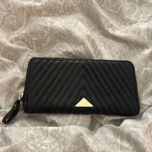 Svart quiltad plånbok med dragkedja - Snygg svart plånbok i quiltad design med dragkedja. Den har flera fack för kort och en praktisk dragkedja i guld för mynt. Perfekt för att hålla ordning på dina saker med stil!