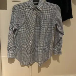 Säljer en klassisk rutig skjorta från Polo Ralph Lauren i blått och vitt. Skjortan har långa ärmar och knappar framtill. Perfekt för både vardag och festliga tillfällen. Den är i mycket bra skick och gjord av bomull för en bekväm passform. Storlek xxs ungefär.