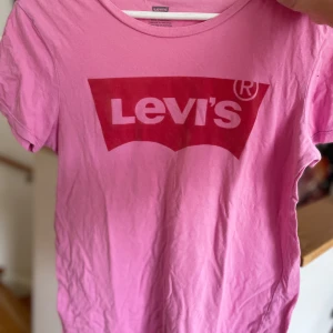 Rosa t-shirt från Levi's - Säljer en snygg rosa t-shirt från Levi's i storlek S. Den har en klassisk rund hals och korta ärmar. Framsidan pryds av det ikoniska Levi's-logotypen i rött. Perfekt för en avslappnad stil eller vardagsbruk!