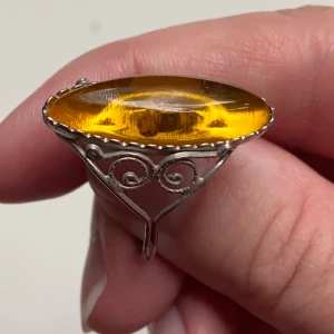 Ring med stor bärnstensfärgad sten - Säljer en unik ring med en stor, oval bärnstensfärgad sten. Stenen är infattad i en detaljerad silverram med vackra snirkliga mönster. Perfekt för att ge en vintagekänsla till din outfit. Passar både till vardags och fest!