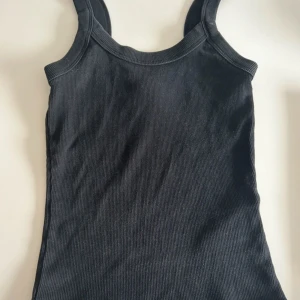 Svart ribbad linnetopp Ginatricot  - Säljer en supersnygg svart ribbad linnetopp i storlek XXS. Använd endast ett fåtal gånger. Perfekt som ett basplagg i garderoben. Toppen har en enkel och klassisk design med breda axelband.