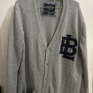 Grå kofta från Bondelid - Säljer en stilren grå kofta från Bondelid i storlek medium. Den har ett stort mörkblått B-broderi på bröstet och bruna armbågslappar. Perfekt för höst och vinter med sin mjuka och bekväma passform. Koftan har knappar framtill och är i mycket bra skick.