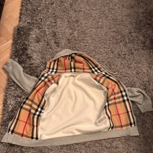 Grå hoodie från Burberry - Säljer en snygg grå hoodie från Burberry med klassiskt rutigt mönster i huvan. Den har dragkedja framtill och är i mycket bra skick. Perfekt för höst och vår, och den är supermjuk och bekväm. Passar både till vardags och lite mer uppklädda tillfällen. 🧥✨stl m men passar S