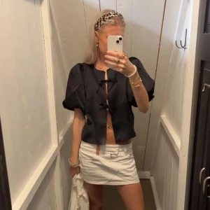 Svart blus med knytning - Snygg svart blus med knytning framtill. Den har korta ärmar och en chic, lite glansig finish. Perfekt för en utekväll eller fest! Passar bra med både kjol och byxor. NYPRIS 600