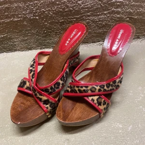 Leopardmönstrade sandaler med träklack - Snygga sandaler med leopardmönster och röda detaljer. De har en träklack och öppen tå, perfekt för sommarens fester. Sandalerna är i bra skick och ger en cool och trendig look.