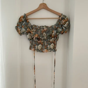Blommig offshoulder topp - Säljer en superfin blommig offshoulder topp med puffiga ärmar. Den har ett smockat liv och knytband framtill. Perfekt för sommardagar eller en festlig kväll. Färgerna är en mix av grönt, orange och vitt. Toppen är croppad och ger en romantisk look.