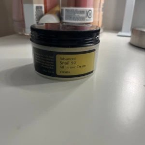 COSRX Advanced Snail 92 All in One Cream - En återfuktande kräm från COSRX med snigelextrakt som ger huden näring och lyster. Perfekt för att återställa hudens elasticitet och mjukhet. Kommer i en praktisk burk med svart lock och gul etikett.