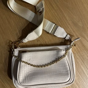 Beige axelväska - Säljer en snygg beige axelväska med krokodilmönster och guldkedja. Väskan har en justerbar axelrem i textil och dragkedja upptill. Har en ficka framtid och rymmer ändå mycket! 