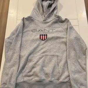 Säljer en snygg grå hoodie från GANT med deras klassiska logga och ett coolt emblem på bröstet. Tröjan är perfekt skick och storleken är 176! Den har en stor ficka fram och en bekväm huva. Perfekt för både vardag och chill! 🩶