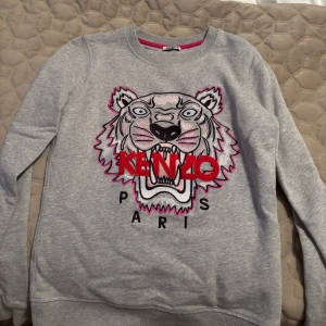 Grå sweatshirt från Kenzo - Säljer en grå sweatshirt från Kenzo med ett coolt tigertryck och texten 'Kenzo Paris' i rött och svart. 