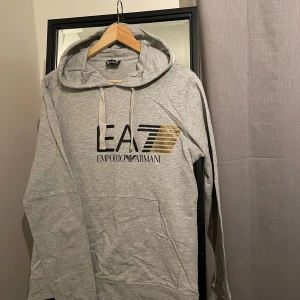 Grå hoodie från Emporio Armani - Säljer en snygg grå hoodie från Emporio Armani i storlek S passar även M, med deras ikoniska logga i svart och guld på bröstet. Den är i bomull och har en bekväm passform med långa ärmar och en praktisk huva med dragsko. Perfekt för både vardag och chill.