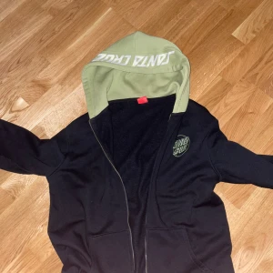 Svart och grön hoodie från Santa Cruz - Säljer en snygg svart hoodie från Santa Cruz med grön insida på huvan och vit text. Den har en dragkedja framtill och är perfekt för både vardag och chill. Märket är tryckt på bröstet och huvan. Passar perfekt för höst och vår!