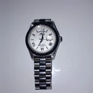 Silverfärgad klocka från Lotus - Ny pris 1600kr. Säljer en stilren silverfärgad klocka från Lotus. Den har en vit urtavla med romerska siffror och dag/datumvisning. Armbandet är i rostfritt stål och har en klassisk design. Perfekt för både vardag och fest!
