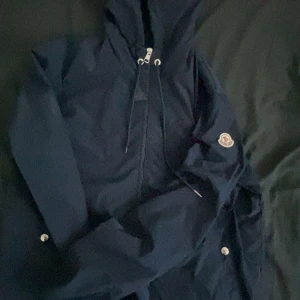Mörkblå vindjacka med huva - Säljer en snygg mörkblå vindjacka från Moncler. Den har en praktisk huva och dragkedja framtill. Perfekt för blåsiga dagar och har en stilren design med Moncler-loggan på ärmen. Passar både till vardags och lite finare tillfällen. Ny pris för denna e ungefär 5500 jag själv som andvänt den är villig att gå ner till 2000 pris kan diskuteras. Har andvänts i 4 månader. Den är köpt från trendtorget