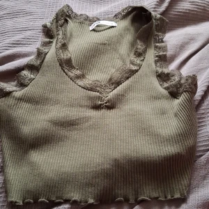 Olivgrön ribbad topp med spetsdetaljer - Säljer en superfin olivgrön ribbad topp från new yorker. Den är ärmlös och har v-ringning med spets detaljer runt halsen och ärmarna. Perfekt för sommardagar eller som ett lager under en jacka. Toppen är croppad och ger en snygg look till både jeans och kjol.