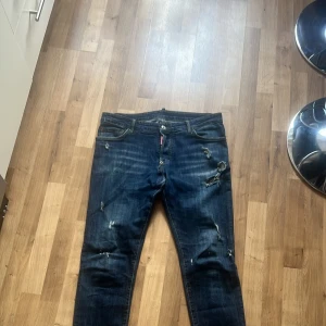 Blå jeans från Dsquared2 - Säljer ett par snygga blå jeans från Dsquared2 med slitna detaljer och en cool DD-logga på bakfickan. De har en knappgylf och är i ett klassiskt jeansmaterial. Hör av er vid intresse!(finns inget kvitto eller så) 