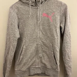 Grå hoodie från Puma - Säljer en grå hoodie från Puma i bra skick. Den har en dragkedja framtill och en rosa Puma-logga på bröstet. Perfekt för höst och vår, med långa ärmar och en skön huva. Passar både till vardags och träning.